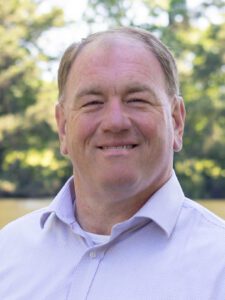 Professional headshot of Joseph N. Hocutt II, J.D., in a light-colored dress shirt, standing outdoors in front of trees and a lake.
