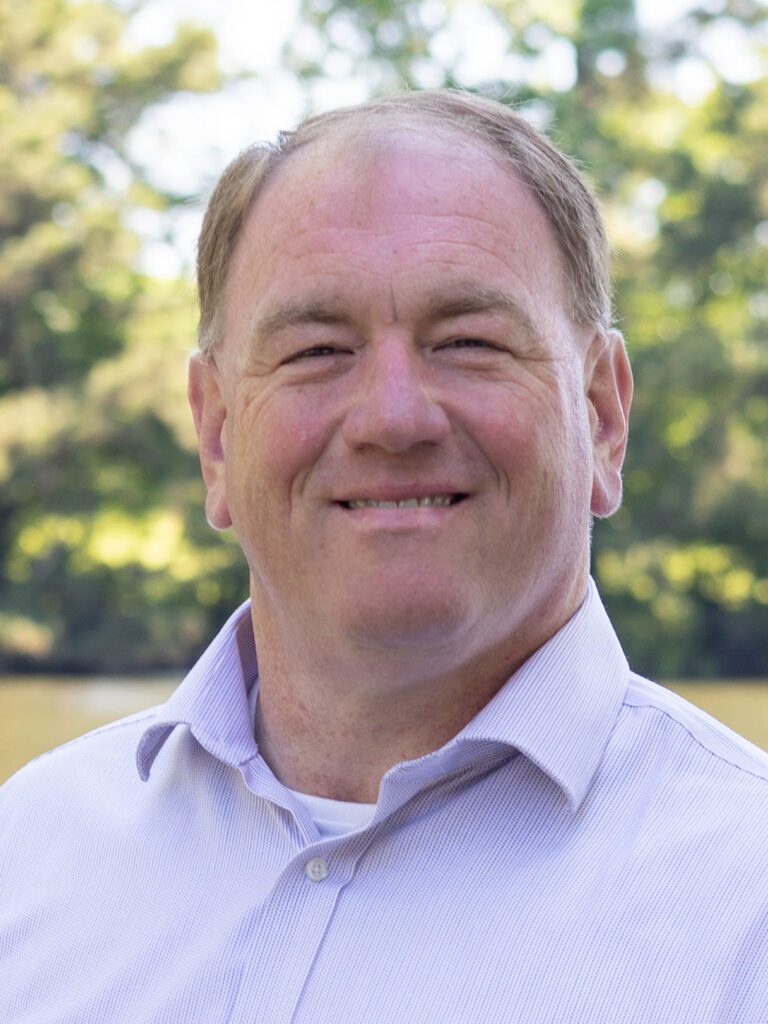 Professional headshot of Joseph N. Hocutt II, J.D., in a light-colored dress shirt, standing outdoors in front of trees and a lake.