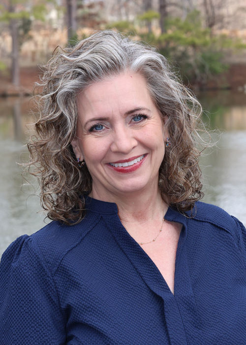 Smiling woman with curly gray hair in blue blouse, outdoors by a lake.