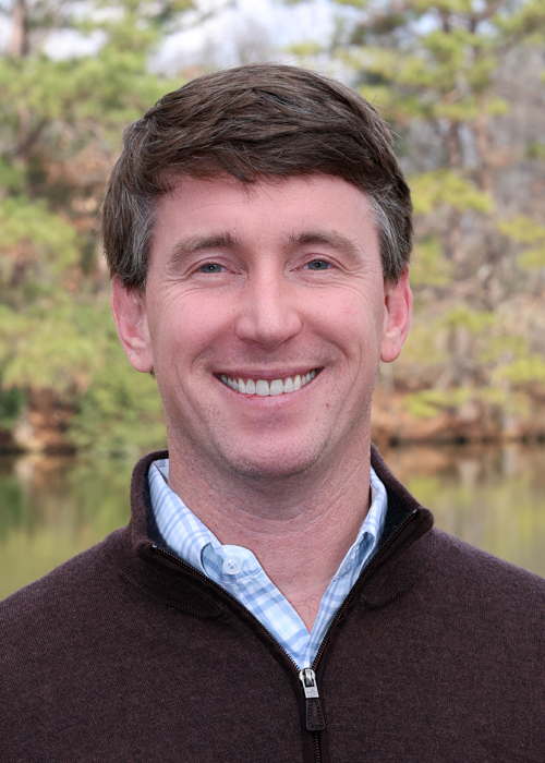 Smiling man in a brown sweater, outdoors with a lake background