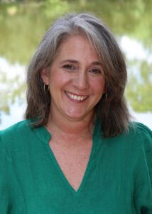 Smiling woman with gray hair in a green blouse stands outdoors by water.