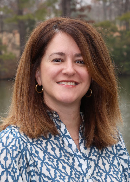 Portrait of a smiling woman with brown hair wearing a blue and white patterned shirt.