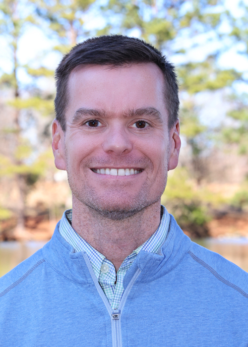 Headshot of a smiling man in a blue sweater and collared shirt outdoors.